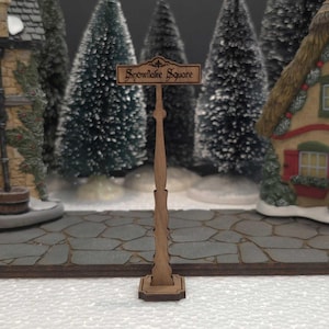 May include: A wooden signpost with a brown wooden sign that reads "Snowflake Square". The signpost is standing on a stone path in front of two miniature houses.