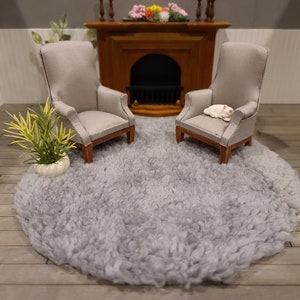 May include: Two gray upholstered armchairs with wooden legs, one with a white cat figurine sitting on the seat, on a gray fluffy round rug in a room with a fireplace.