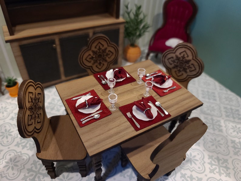 May include: A miniature wooden dining table set with four chairs, each with a carved snowflake design. The table is set with four place settings, each with a red napkin, a white plate, and silver cutlery. There are also four wine glasses on the table.