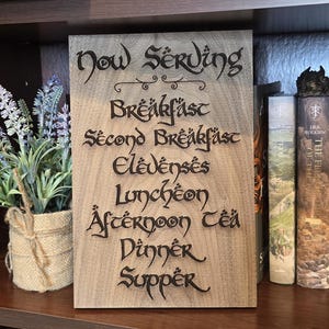 May include: Wooden sign with the words "Now Serving" and meal times in decorative lettering. A small potted lavender plant and several books are on a wooden shelf. A dragon figurine sits atop the books.