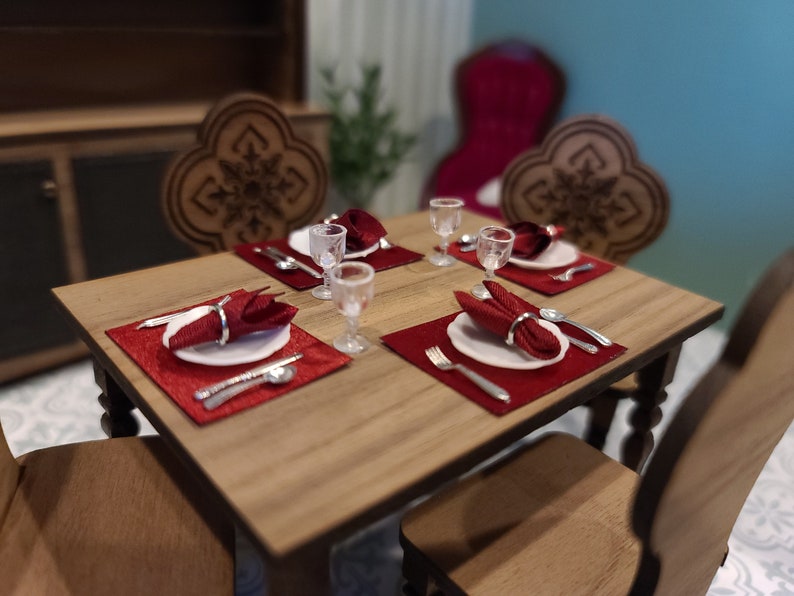 May include: A wooden dining table set with four place settings. Each place setting includes a white plate, a red napkin, silverware, and a clear glass. The table has a wood grain finish and is surrounded by four chairs.