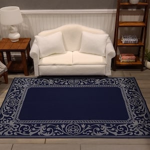 May include: A navy blue miniature rug with a gray floral and scrollwork border. The rectangular rug is placed in a dollhouse living room setting, with a white sofa, side table, and lamp visible in the background.