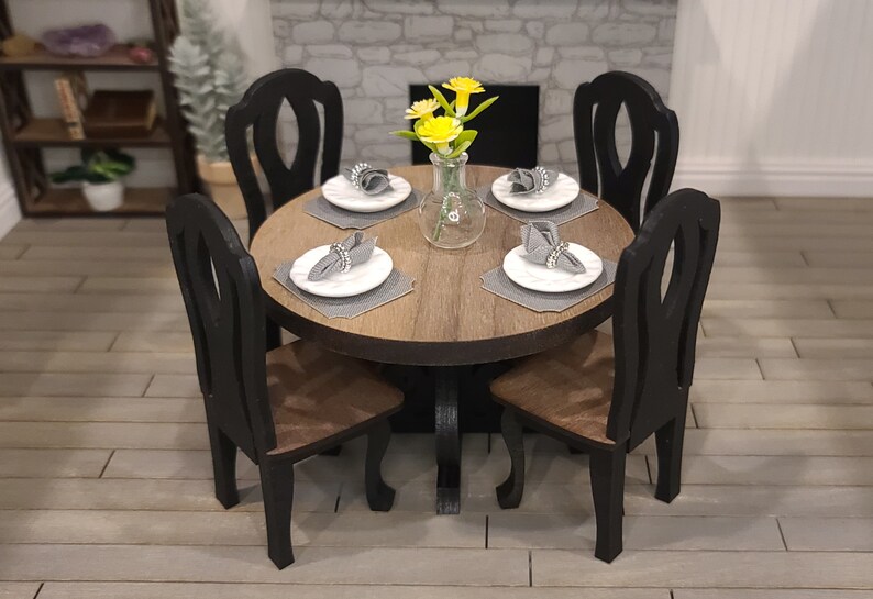 Peut inclure: Une table ronde en bois avec quatre chaises noires. La table est dress&eacute;e avec quatre couverts, chacun avec une assiette blanche, une serviette grise et un anneau de serviette argent&eacute;. Un vase avec des fleurs jaunes se trouve au centre de la table.