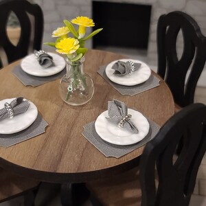 Peut inclure: Une table ronde en bois avec quatre chaises noires. La table est dress&eacute;e avec quatre couverts, chacun avec une assiette blanche, une serviette grise et un anneau de serviette argent&eacute;. Un vase avec des fleurs jaunes se trouve au centre de la table.