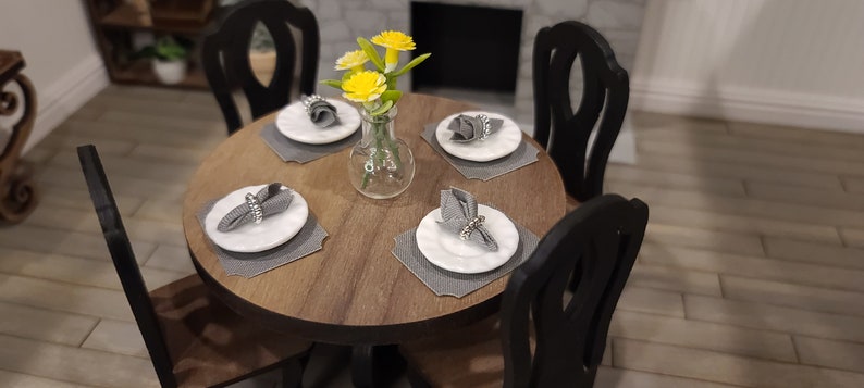 Peut inclure: Une table ronde en bois avec quatre chaises noires. La table est dress&eacute;e avec quatre couverts, chacun avec une assiette blanche, une serviette grise et un anneau de serviette argent&eacute;. Un vase avec des fleurs jaunes se trouve au centre de la table.