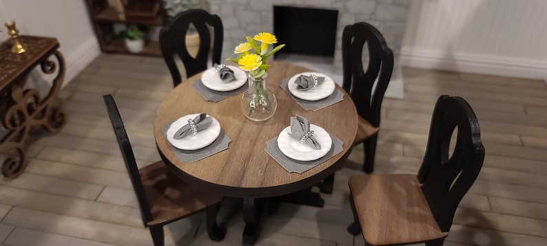 Peut inclure: Une table ronde en bois avec quatre chaises noires. La table est dress&eacute;e avec quatre couverts, chacun avec une assiette blanche, une serviette grise et un anneau de serviette argent&eacute;. Un vase avec des fleurs jaunes se trouve au centre de la table.
