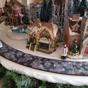 May include: A miniature village scene with a bakery, a church, and a horse-drawn carriage. The scene is set on a white surface with a gray stone path.
