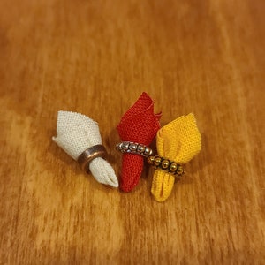 Puede incluir: Tres servilletas de tela con anillos decorativos. Las servilletas son blancas, rojas y amarillas. Los anillos son plateados y dorados.