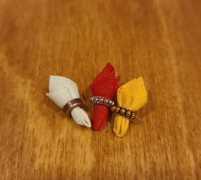 Puede incluir: Tres servilletas de tela con anillos decorativos. Las servilletas son blancas, rojas y amarillas. Los anillos son plateados y dorados.