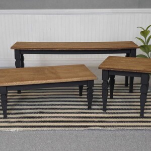 May include: Three wooden tables with black painted legs and a natural wood top. The tables are arranged in a row from largest to smallest, with the largest table at the back and the smallest table at the front. The tables are on a striped rug.
