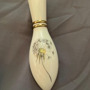 May include: A cream-colored ceramic bud vase with a tapered, elongated shape. The neck features two thin, horizontal gold-toned bands. The body is decorated with a detailed black and yellow line drawing of a dandelion seed head in full bloom.