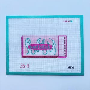 May include: A needlepoint canvas with a teal border and a light blue background. The canvas features a pink rectangle with the words "bad bitch" in teal, surrounded by teal flames. The numbers "55-11" and "s/s" are also visible.