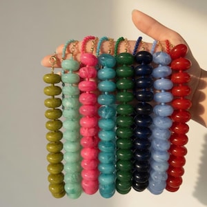 May include: A collection of eight necklaces featuring round, disc-shaped beads in various marbled colors including olive green, mint, pink, turquoise, forest green, navy blue, light blue, and red, each attached to a gold-toned metal chain.