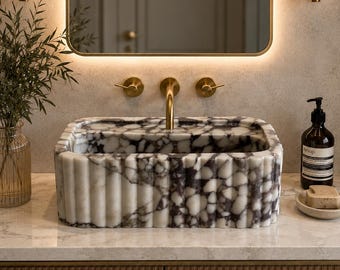 May include: A rectangular marble sink with a fluted exterior and a white and dark purple veined pattern. The sink features a brass faucet with two matching handles, positioned below a rectangular mirror with integrated LED backlighting.
