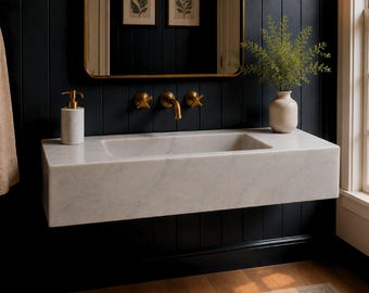 May include: A wall-mounted white marble rectangular sink with a brass faucet and cross-handle controls. The sink is positioned against a black vertical paneled wall featuring a gold-framed mirror, two brass wall sconces, and a marble soap dispenser.
