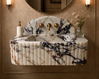 May include: A wall-mounted bathroom sink crafted from white marble with prominent dark purple and brown veining. The basin features a fluted exterior design and a decorative arched backsplash. It includes brass fixtures and a soap dispenser.
