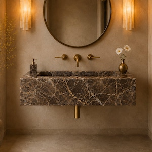 May include: A wall-mounted rectangular brown marble sink with white veining, featuring a brass faucet and two handles. Above the sink is a round mirror with a dark frame, flanked by two vertical wall sconces emitting a warm, soft golden light.
