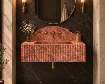 May include: A wall-mounted pink marble sink featuring a fluted front design, a matching backsplash, and gold-toned hardware. The sink is positioned beneath a round gold-framed mirror flanked by two cylindrical wall sconces with warm lighting.