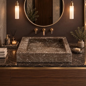 May include: A rectangular brown marble sink sits on a matching marble countertop with a dark wood vanity cabinet. Above the sink is a round mirror with a gold frame, flanked by two cylindrical pendant lights and gold wall-mounted faucets.