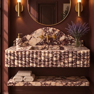 May include: A wall-mounted bathroom vanity featuring a fluted front and a backsplash made of white and deep burgundy marble. The unit includes a matching marble shelf below, a gold faucet, a soap dispenser, and a round mirror with a gold frame.