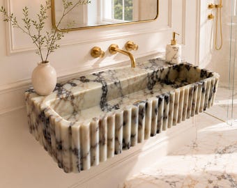 May include: A wall-mounted rectangular bathroom sink crafted from white marble with prominent grey and black veining. The exterior features a vertical fluted design. A gold faucet is mounted on the wall above the basin, accompanied by a matching marble soap dispenser and a white vase with green branches.