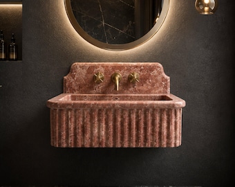 May include: A wall-mounted rectangular sink crafted from pink marble with a fluted front design. The sink features a high backsplash and is equipped with a central brass faucet and two matching brass cross-handle knobs for water control.