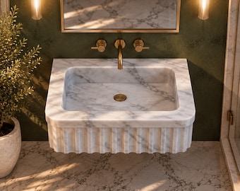 May include: A wall-mounted white marble sink featuring a square basin with a fluted exterior design. The sink includes a gold-toned faucet and two matching gold-toned handles mounted on a dark green wall beneath a rectangular mirror.
