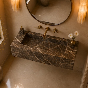 May include: A wall-mounted rectangular bathroom sink crafted from dark brown marble with prominent white veining. The sink features a brass faucet with two matching handles, a soap dispenser, and a small brass vase holding two white flowers.