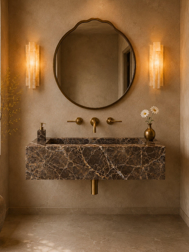 May include: A wall-mounted rectangular brown marble sink with white veining, featuring a brass faucet and two handles. Above the sink is a round mirror with a dark frame, flanked by two vertical wall sconces emitting a warm, soft golden light.