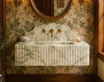 May include: A wall-mounted white and green marble sink featuring a fluted front design, a scalloped backsplash, and brass fixtures. The sink is positioned beneath a large oval mirror with a gold frame and two brass wall sconces with glass globes.
