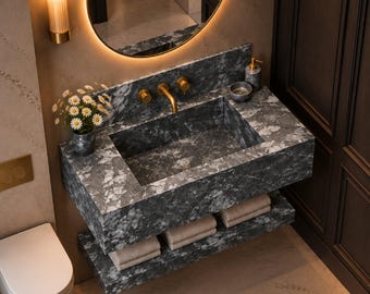 May include: A wall-mounted bathroom vanity crafted from dark gray marble with white veining, featuring a rectangular basin, a gold-toned faucet, and two matching shelves below holding folded beige towels. A small vase with white flowers sits nearby.
