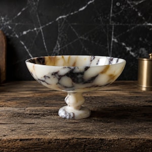 May include: A footed bowl crafted from polished stone featuring a creamy white base color with prominent dark brown and black veining throughout the surface. The bowl has a wide, shallow basin supported by a thick, rounded pedestal base.
