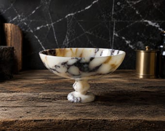 May include: A footed bowl crafted from polished stone featuring a creamy white base color with prominent dark brown and black veining throughout the surface. The bowl has a wide, shallow basin supported by a thick, rounded pedestal base.