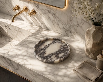 May include: A circular marble tray featuring a white and dark gray veined pattern. The tray has a smooth, polished surface with a raised edge, resting on a matching white and gray marble countertop next to a textured vase and linen cloth.