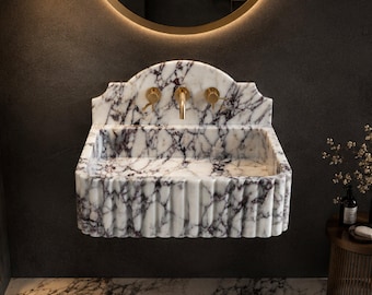 May include: A wall-mounted bathroom sink crafted from white marble with prominent dark purple veining. The basin features a fluted exterior design and an arched backsplash. It is equipped with three gold-toned metal fixtures, including a central faucet.