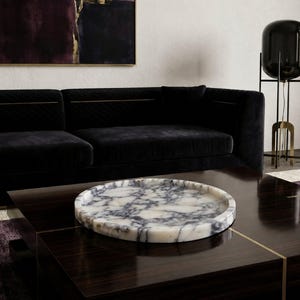 May include: A circular decorative tray crafted from white marble with prominent dark grey and black veining. The tray features a smooth, polished surface and a raised, rounded edge, presenting a natural stone pattern throughout its entire structure.