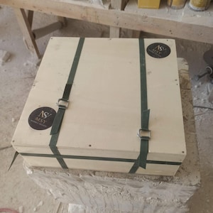 May include: A square light-colored wooden crate secured with two dark green nylon straps and metal buckles. Two circular black stickers with the white text MSY MARBLE DESIGN are affixed to the top surface of the wooden box.