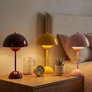 Peut inclure: Trois lampes de table avec des abat-jours en forme de dôme, de couleur bordeaux, jaune et rose. Un petit cactus en pot beige, un pichet beige et une horloge dorée sont sur la table de chevet en bois. Les lampes mesurent environ 30 cm de haut.