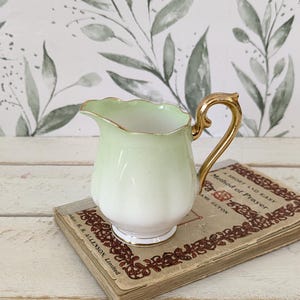 May include: A porcelain creamer featuring a pale green gradient finish, a scalloped rim, and a gold-toned handle. The creamer rests on a vintage book titled A Short and Easy Method of Prayer by Madame Guyon, published by H. R. Allenson, Limited.