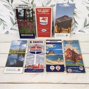 May include: A collection of seven vintage paper travel maps and guides featuring various US states including Arizona, Nevada, Utah, New Jersey, Delaware, Idaho, and North Dakota, with colorful illustrations, brand logos like Mobilgas and Standard, and text.