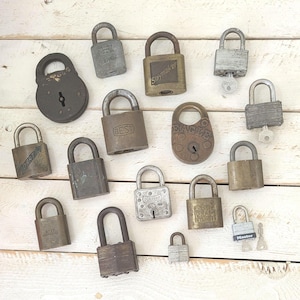 May include: A collection of vintage padlocks in various sizes and shapes, arranged on a light-colored wooden surface. The padlocks are made of metal and show signs of wear, with some having keys. Some padlocks have text such as "BEST" and "Master".