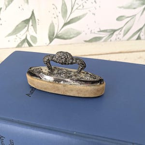 May include: A small, antique silver-toned iron with a floral handle and intricate detailing. The iron rests on a tan, cushioned base. The iron is displayed on a blue book.