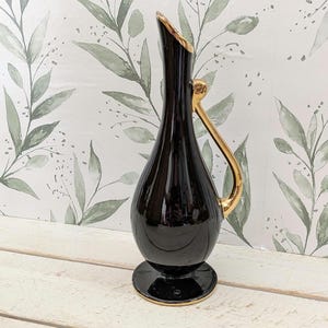 May include: A glossy black ceramic vase with a teardrop-shaped body, a slender neck, and a circular base. The vase features a metallic gold handle extending from the neck to the body, with gold trim along the rim and the edge of the base.