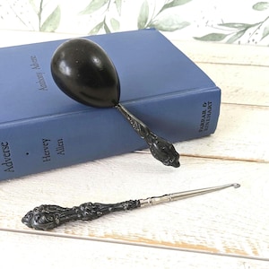 May include: A black, egg-shaped spoon and a crochet hook with ornate, dark metal handles. The spoon rests on a blue book titled "Adverse" by Hervey Allen. The crochet hook lies below, on a white wooden surface.