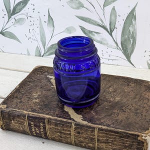 May include: A small, cylindrical cobalt blue glass jar with a threaded neck and embossed lettering around the upper shoulder. The jar features a barrel-like shape with a wide opening and a smooth, translucent finish, resting on a weathered, dark brown antique book.