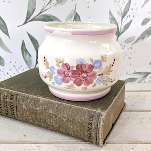 May include: A white ceramic pot with a pink rim and base, adorned with floral designs in shades of pink, burgundy, and blue. The pot sits atop a vintage book with the text "Heath & Co." on the spine. The background features a watercolor floral wallpaper.