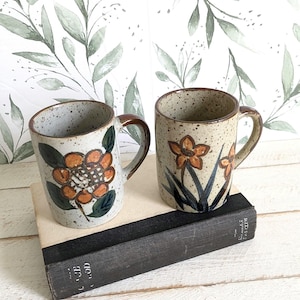 May include: Two ceramic mugs with a speckled light gray finish. The left mug features a large orange flower with dark green leaves. The right mug displays two orange flowers with long dark green stems and leaves. Both mugs have brown handles.