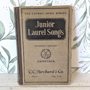 May include: A vintage book titled "Junior Laurel Songs" from "The Laurel Music Series." The cover is tan with a black rectangular label featuring the title and author, "Armitage." The publisher is C.C. Birchard & Co., with locations in Boston and New York.