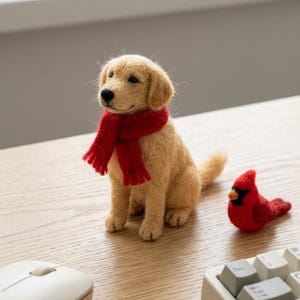 May include: A needle-felted golden retriever figurine with a bright red scarf wrapped around its neck, positioned next to a small, needle-felted red cardinal bird figurine on a light-coloured wooden surface.