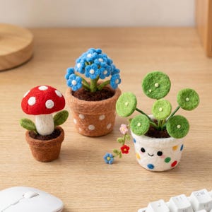Könnte beinhalten: Drei Miniatur-Filzpflanzen in Terrakotta-Töpfen. Ein roter Pilz mit weißen Punkten, eine blaue Blume und eine grüne Pflanze mit runden Blättern. Die Töpfe haben weiße Punkte und ein lächelndes Gesicht.